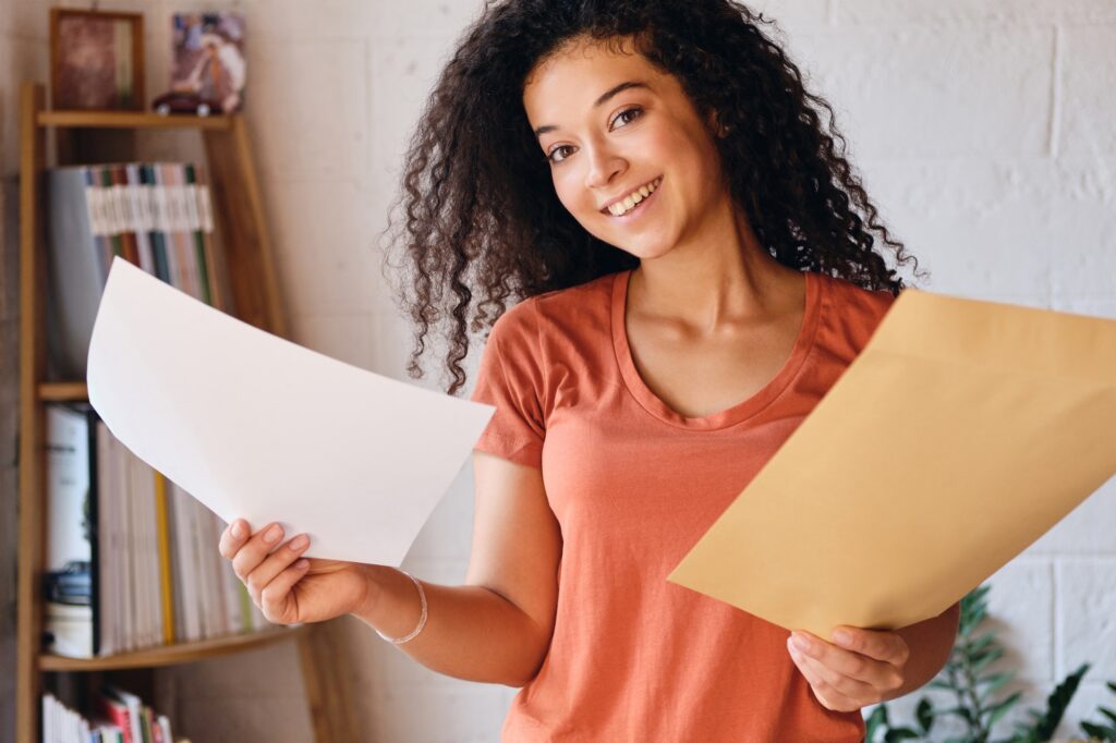 Young cheerful woman in T-shirt joyfully looking in camera holding letter with exam results in hands